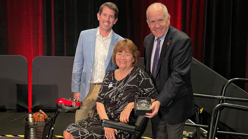 Board Chair Hampton Graham, CEO Ray Carson and Award recipient Molly Mahoney smile at the camera as she is being presented with a crystal plaque.